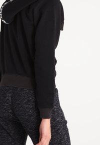 Felpa nera con fodera a fantasia, caratterizzata da orlo rotondo e polsini a costine. Abbinata a pantaloni sportivi scuri e strutturati.