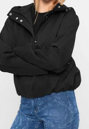 Person iført en sort hættetrøje med sølvknapper og blå mønstrede jeans, arme krydset over brystet.
