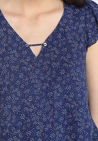 Primo piano di una blusa blu navy con motivo floreale bianco e rosso, caratterizzata da un scollo a V fissato da una piccola fascia con due bottoni.