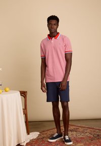 Rood en wit gestreepte polo shirt met een rode kraag en blauwe accenten, gecombineerd met marineblauwe shorts en donkere sneakers. Eenvoudig, casual ontwerp.