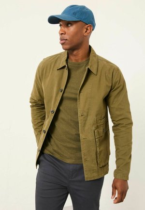 Olivgrüne Baumwolljacke mit Knopfverschluss und Seitentaschen, getragen über einem passenden grünen Hemd. Blaue Mütze, dunkle Hose.