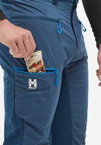 Main tenant une barre énergie étiquetée "BAOUW Extra" dans la poche de cuisse zippée bleue d'un pantalon de randonnée Millet.