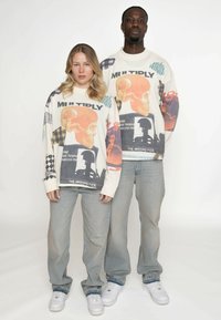 Zwei Personen tragen übergroße beigen Sweatshirts mit grafischen Drucken in lebhaften Farben, kombiniert mit hellblauen Jeans und weißen Turnschuhen.
