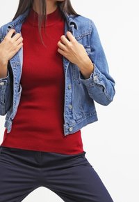 Vrouw in een rode geribbelde coltrui, blauwe denim jas en marineblauwe broek, die de kraag van haar jas met beide handen aanpast.