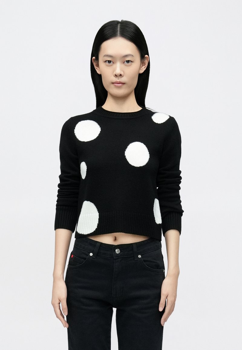 Suéter negro corto con grandes lunares blancos, cuello y puños acanalados, combinado con jeans negros con cierre de botón y detalles de bolsillo.