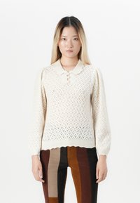 Junge Frau mit langen Ombre-Haaren trägt einen cremefarbigen Strickpullover mit Knöpfen und puffigen Ärmeln, kombiniert mit braunen und schwarzen vertikal gestreiften Hosen.