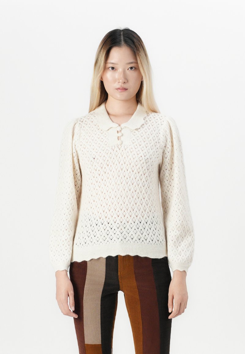 Junge Frau mit langen Ombre-Haaren trägt einen cremefarbigen Strickpullover mit Knöpfen und puffigen Ärmeln, kombiniert mit braunen und schwarzen vertikal gestreiften Hosen.