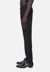 Man som bär slim svarta jeans och svarta läder slip-on skor med ankelremmar, stående på sidan mot en ljus vit bakgrund.