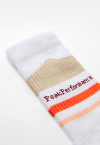 Bijela atletska čarapa s teksturiranim bež dijelom, narančastim i breskvastim prugama, te "PeekPerformance" logom u tamnocrvenoj boji.