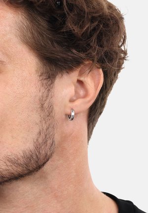 Orecchino a cerchio in argento indossato nel lobo, con una texture lucida e un design circolare semplice. I capelli sono ricci e la barba è corta.