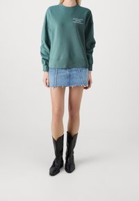 Felpa verde con colletto rotondo e logo ricamato, abbinata a una mini gonna in denim strappata e stivali cowboy neri in pelle.
