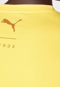 Žluté sportovní tričko s hladkým, elastickým materiálem. Na zadní straně se nachází hnědé logo a text. Jednoduchý design s klasickým kulatým výstřihem.