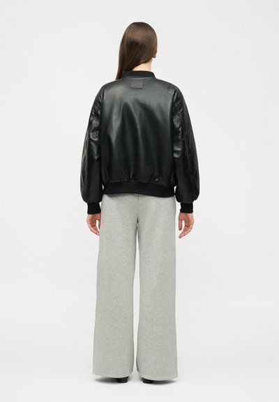 Žena s dlhými hnedými vlasmi, oblečená v čiernej koženej bomber bunde a širokých sivých nohaviciach, stojaca chrbtom k bielej stene.