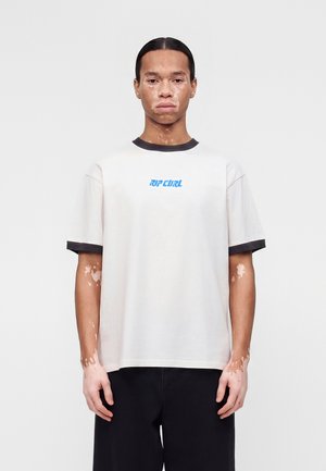 Άνδρας με λεύκη που φοράει ένα λευκό t-shirt Rip Curl με μαύρο περίγραμμα και μαύρο παντελόνι, στέκεται μπροστά σε ένα απλό ανοιχτό φόντο.