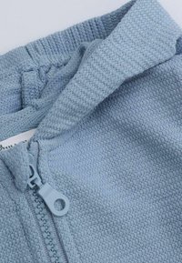 Jasnoniebieska bluza z zamkiem, teksturowany materiał z prążkowanym kołnierzykiem, z gładkim zamkiem błyskawicznym i widoczn ą metką przy dekolcie.