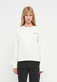 Kremowy sweatshirt o lekko oversizedowym kroju, okrągłym dekolcie i haftowanym napisie "Hawaii" w niebieskim kolorze na lewej piersi. Miękki materiał bawełniany.