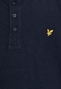 Navy polo paita valmistettu pehmeästä kankaasta, jossa on kolmen napin avaus, brodeerattu keltainen logo ja teksturoitu kauluksen yksityiskohta.