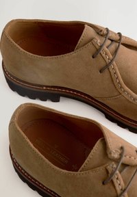 Chaussures en suede marron avec lacets, bout rond, surpiqûres blanches contrastées et semelle en caoutchouc noir texturé. L'intérieur révèle une doublure en cuir.