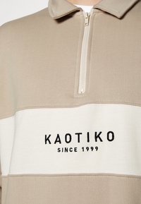 Beige och krämfärgad sweatshirt med dragkedja vid kragen. Har "KAOTIKO" och "SINCE 1999" broderat i svart på framsidan, mjuk textur.