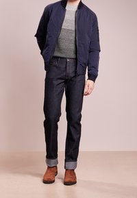 Homme portant une veste bomber bleu marine, un pull en tricot gris, un jean foncé à revers et des bottes en suède marron, se tenant contre un fond beige.