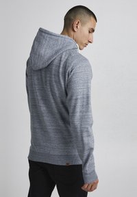 Sudadera con capucha gris de tejido texturizado y jaspeado. Presenta mangas largas, puños acanalados y un pequeño parche con logo cerca del dobladillo.