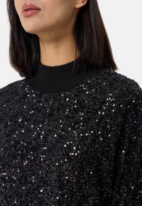 Maglione nero con paillettes, dalla superficie testurizzata e lucida; presenta un colletto a costine nero. La vista ravvicinata mette in evidenza il dettagliato motivo di paillettes.