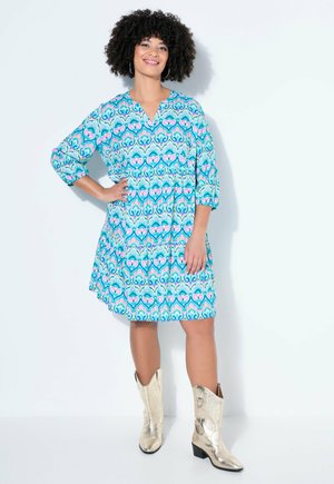 Femme debout, main sur la hanche, portant une robe mi-longue à motifs bleus, roses et blancs, et des bottes de cow-boy argentées métalliques.