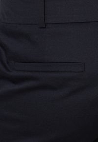 Poche arrière d'un pantalon bleu foncé sur mesure, de style passepoilé, avec une passants de ceinture au-dessus de la poche.