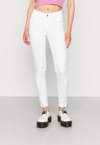 Jeans ajustados blancos con cierre de botón, bolsillos frontales y cremalleras en los tobillos. Combinados con zapatos blancos con cordones negros y suelas gruesas.