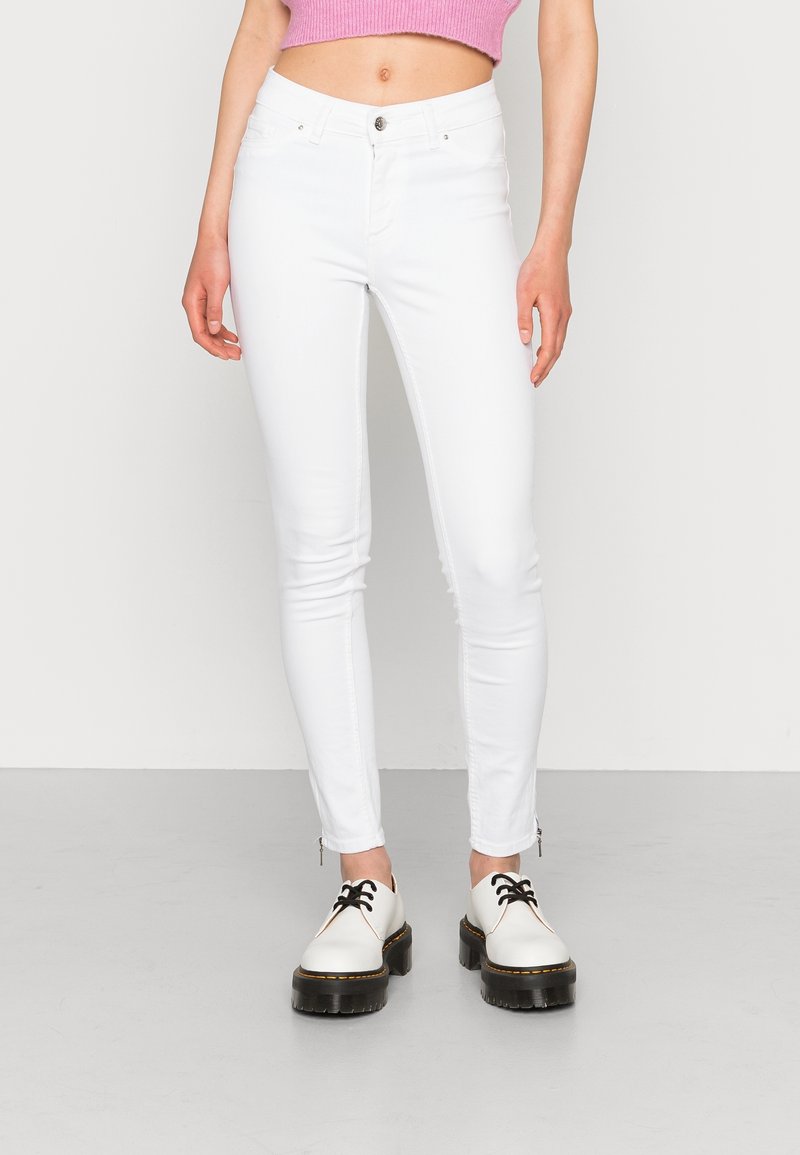 Jeans ajustados blancos con cierre de botón, bolsillos frontales y cremalleras en los tobillos. Combinados con zapatos blancos con cordones negros y suelas gruesas.