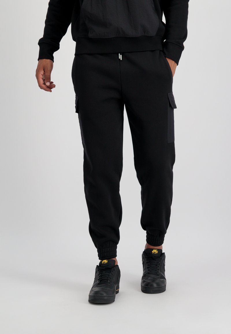 Schwarze Sweatpants aus strukturiertem Stoff mit Cargo-Taschen, elastischen Bündchen und lockerem Schnitt. Kombiniert mit schwarzen Sneakern.