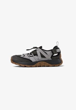 Scarpa sportiva con tomaia in tessuto nero e grigio, suola in gomma, strappi elastici e un'etichetta per facilitarne l'indossamento. Presenta una suola esterna con grip testurizzato.