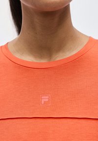 Λαιμός και άνω μέρος στήθους γυναίκας που φοράει πορτοκαλί πουκάμισο με μικρό, κεντρικά τοποθετημένο τετράγωνο λογότυπο με ένα στυλιζαρισμένο "F" στο ύφασμα.