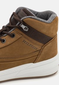 Brązowe skórzane sneakersy typu high-top z ciemnobrązowymi akcentami, teksturowanym szarym kołnierzem, czarnymi elementami metalowymi i białą gumową podeszwą. Posiadają haftowane logo.