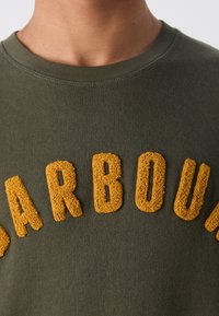 Gros plan sur une personne portant une chemise vert olive avec des lettres texturées jaune moutarde formant le mot "SARBOUN" sur la poitrine.