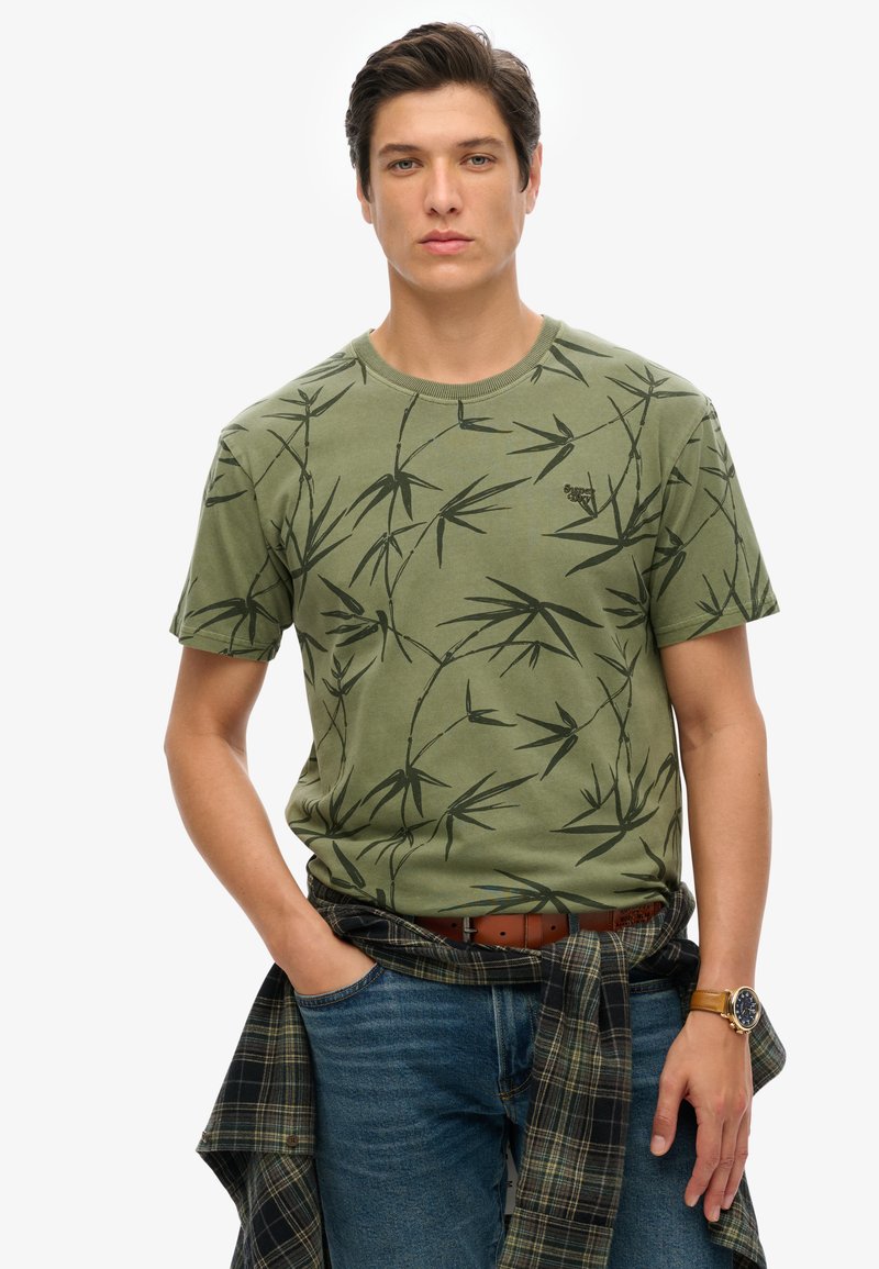 Camiseta de algodón verde con patrón de hojas de bambú negras, cuello redondo, mangas cortas, usada con una camisa de cuadros azul atada a la cintura.