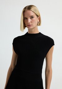 Vestito nero a coste con collo alto e maniche a cappuccio, caratterizzato da un design testurizzato e una vita aderente che si trasforma in una gonna svasata.