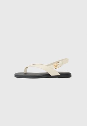 Beige flad sandal med sort sål, der har en tå-rem og en justerbar bagrem med en guldlås.