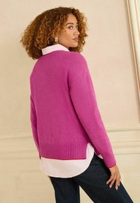 Rózsaszín pulóver bordázott szegéllyel és mandzsettával, fehér, ívelt aljú ing fölött rétegezve. Az anyag puha, sima textúrájú.