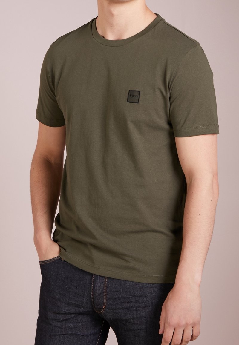 Olijfgroene katoenen t-shirt met korte mouwen, ronde hals en een klein zwart vierkant logo-patch op de borst. Draagt met donkere spijkerbroek.