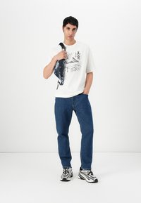 T-shirt graphique blanc avec un design de montagne, jeans en denim bleu, et un sac en bandoulière à motif sombre, porté avec des baskets noir et blanc.