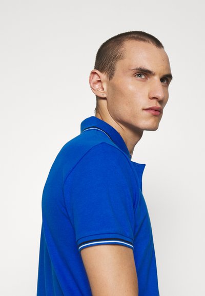 Βασιλικό μπλε πόλο μπλουζάκι με κλασικό γιακά, διαθέτοντας αντίθετους ριγέ τόνους στα μανίκια. Μαλακό ύφασμα με εφαρμοστή σχεδίαση.