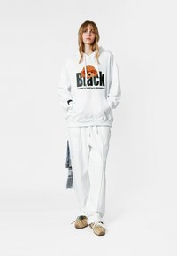 Joven de pie con sudadera blanca con gráfico de perro y pantalones blancos a juego, usando zapatillas beige sobre fondo liso.