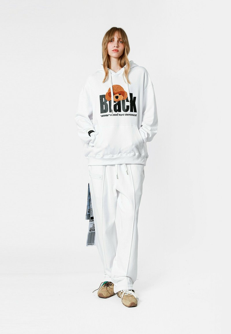 Joven de pie con sudadera blanca con gráfico de perro y pantalones blancos a juego, usando zapatillas beige sobre fondo liso.