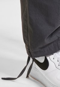 Szare spodnie cargo z ściągaczem na dole oraz białe sneakersy z czarnym logo Nike, skórzana cholewka o fakturze oraz gumowa podeszwa.