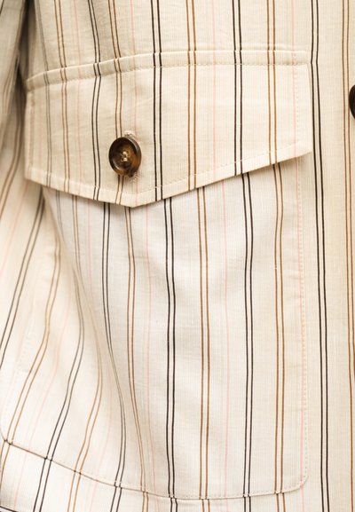 Gros plan d’un tissu rayé beige et blanc avec des lignes marron, noires et roses ; doté d’une poche à rabat avec un bouton marron foncé.