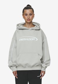 Light gray oversized hoodie with a large white "PEGADOR" logo, front pocket, and drawstring hood. Features white side stripes.