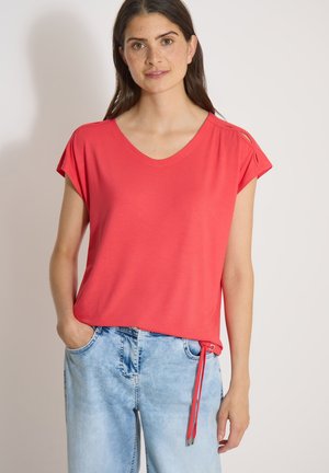 Femme aux longs cheveux bruns portant un top corail à manches courtes et un jean bleu clair, une main dans la poche, debout devant un fond uni.