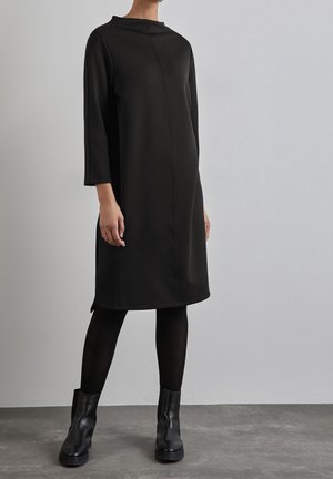 Schwarzes, langärmeliges Kleid mit hohem Ausschnitt, geradem Schnitt, weicher Textur und seitlichen Schlitzen, kombiniert mit schwarzen Knöchel-Stiefeln.