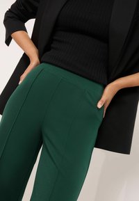 Zaļas piegriezuma bikses ar ribotu melnu augšu, kas izceļas ar gludu tekstūru, asām krokām un pieguļošu jostu, daļēji aizsegta ar melnu bleizeri.